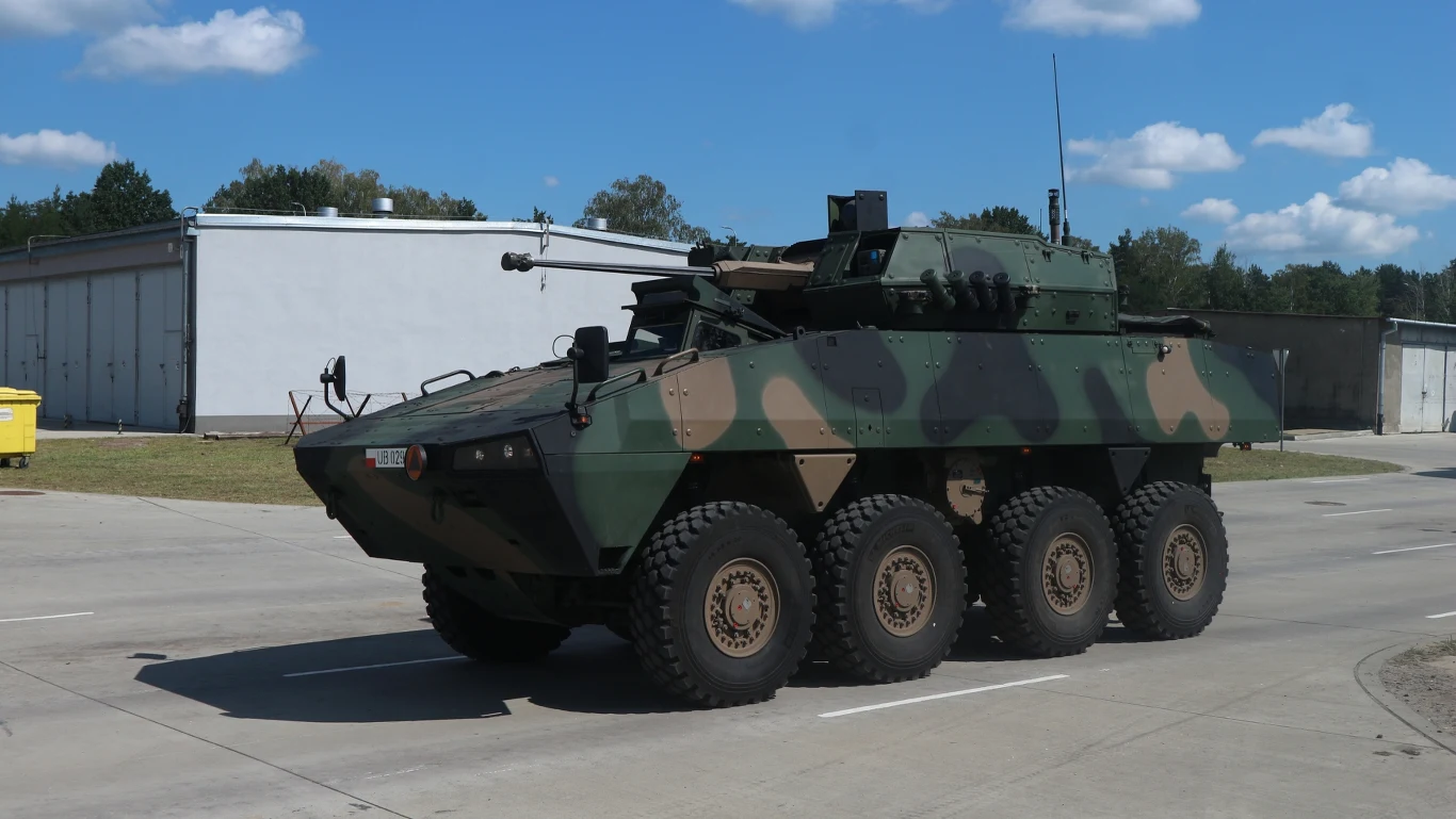 Opancerzony pojazd wojskowy z ośmioma kołami, pomalowany w kamuflaż leśny, ustawiony na betonowej nawierzchni przy budynkach wojskowych, wyposażony w wieżyczkę z działkiem.