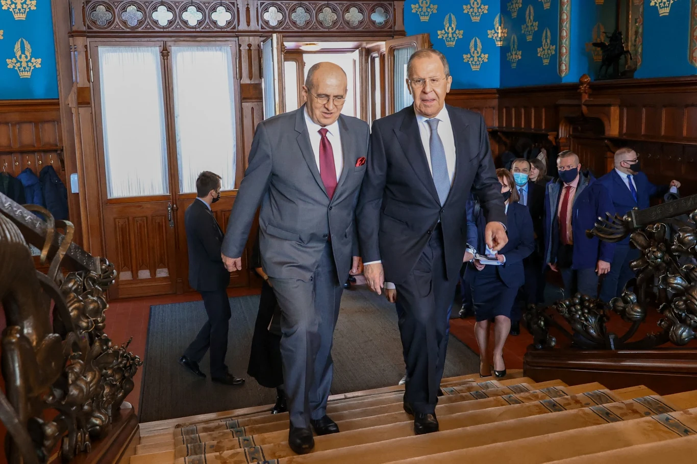 Dwaj mężczyźni w garniturach idą razem po ozdobnych schodach w budynku z bogato zdobionymi drewnianymi balustradami oraz niebieskimi ścianami dekorowanymi złotymi motywami, za nimi podąża grupa innych osób.