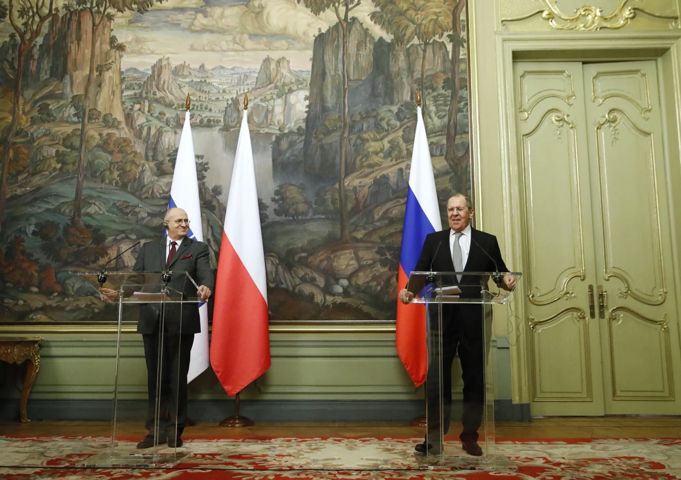 Moskwa, 15 lutego 2022, Zbigniew Rau i Siergiej Ławrow Dwóch mężczyzn w garniturach stoi za przezroczystymi pulpitemi na tle dużych flag Polski i Rosji oraz ozdobnej ściany z malowidłem. Całość otoczona jest eleganckim wnętrzem z orientalnym dywanem i złotymi drzwiami.