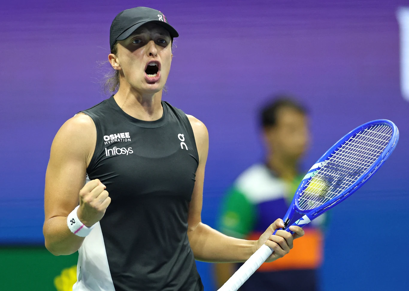 Zawodniczka ubrana w czarną koszulkę sportową i czapkę z daszkiem ściska rakietę tenisową w jednej ręce oraz zaciśniętą pięść w drugiej, wyrażając silne emocje i determinację podczas meczu tenisowego. W tle widoczny rozmazany sędzia lub inny członek ob...