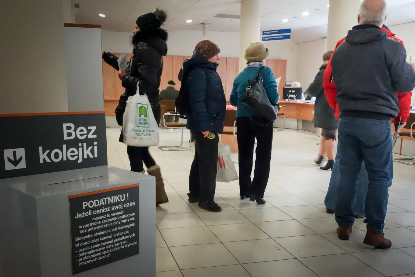 Emeryci odchodzący na emeryturę mogą skorzystać ze zwolnienia podatkowego, aby uniknąć wpadnięcia w drugi próg podatkowy Ludzie stoją w kolejce do stanowiska obsługi w urzędzie, widoczne są krzesła i tabliczki informacyjne, w tle pracownicy za biurkami. Każda osoba ubrana jest w zimową odzież.