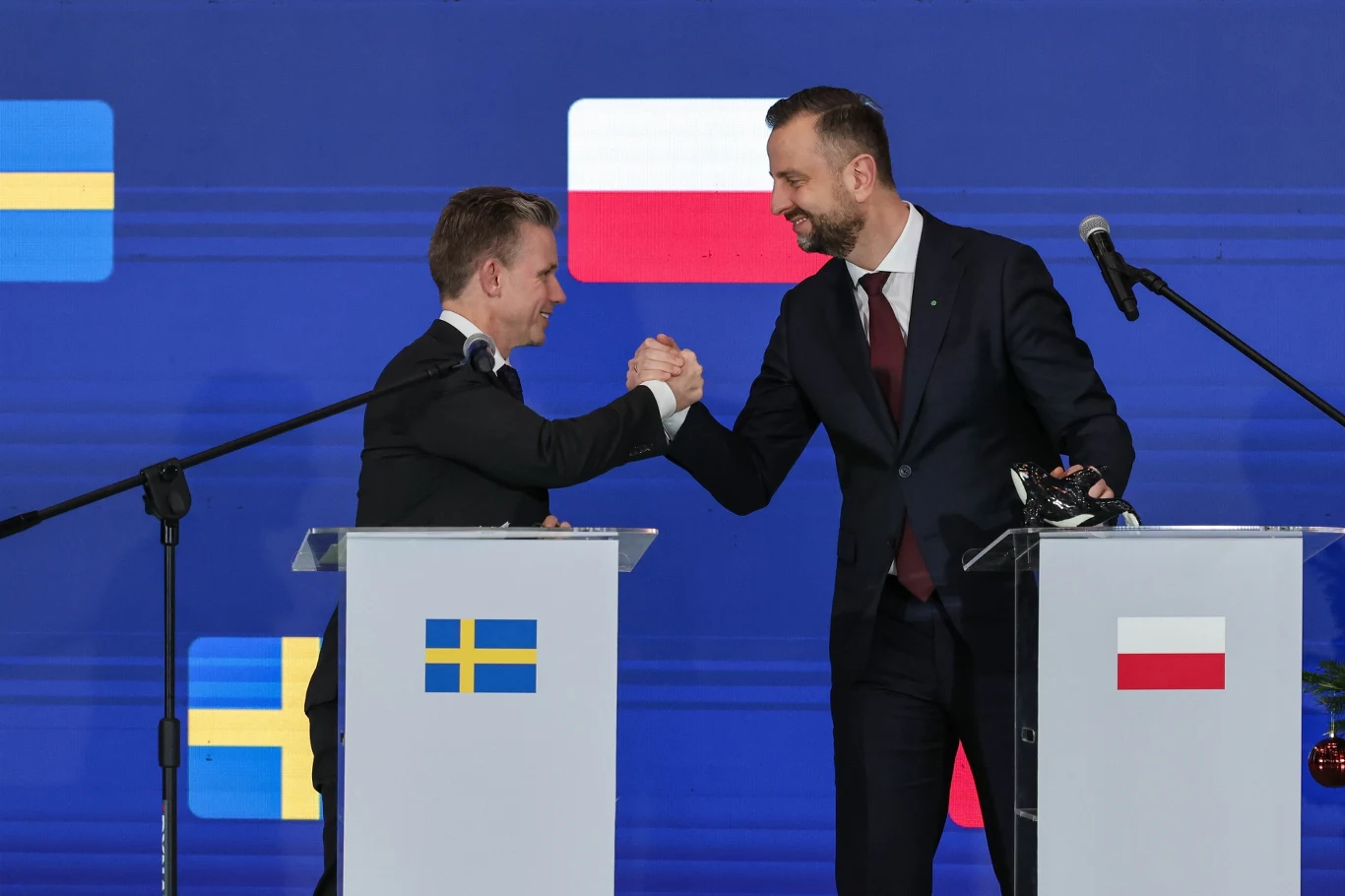 Polska i Szwecja podpisały memorandum ws. okrętów podwodnych Dwóch mężczyzn w garniturach ściska dłonie w geście porozumienia lub współpracy, stojąc przy mównicach oznaczonych flagami Szwecji i Polski na tle niebieskiej ściany z flagami obu państw.