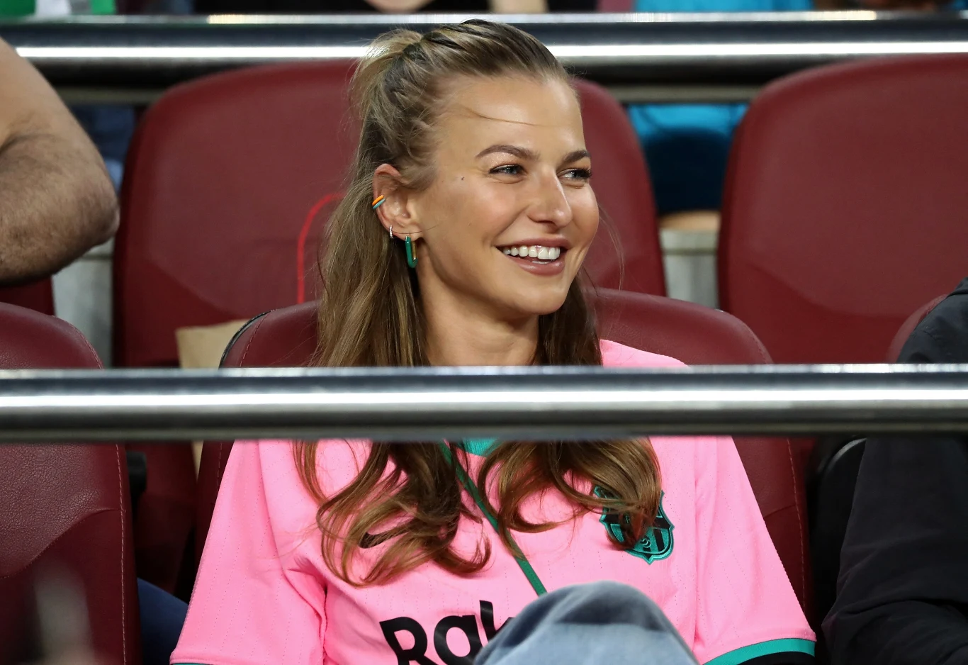 Kobieta o jasnych włosach ubrana w różową koszulkę piłkarską z widocznym logo, siedzi na stadionie na czerwonych krzesłach, uśmiecha się, patrząc w bok.