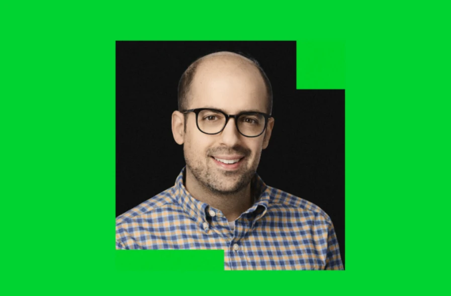 Mężczyzna w okularach, z krótkimi włosami i zarostem, ubrany w koszulę w kratę, uśmiechnięty, na czarnym tle z intensywną zieloną ramką wokół postaci.