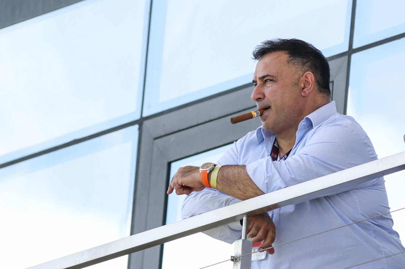 Alex Haditaghi Mężczyzna w białej koszuli stoi na tarasie ze szklaną barierką, oparty o poręcz, trzyma cygaro w ustach, na nadgarstku złoty zegarek, w tle duże przeszklenia budynku.