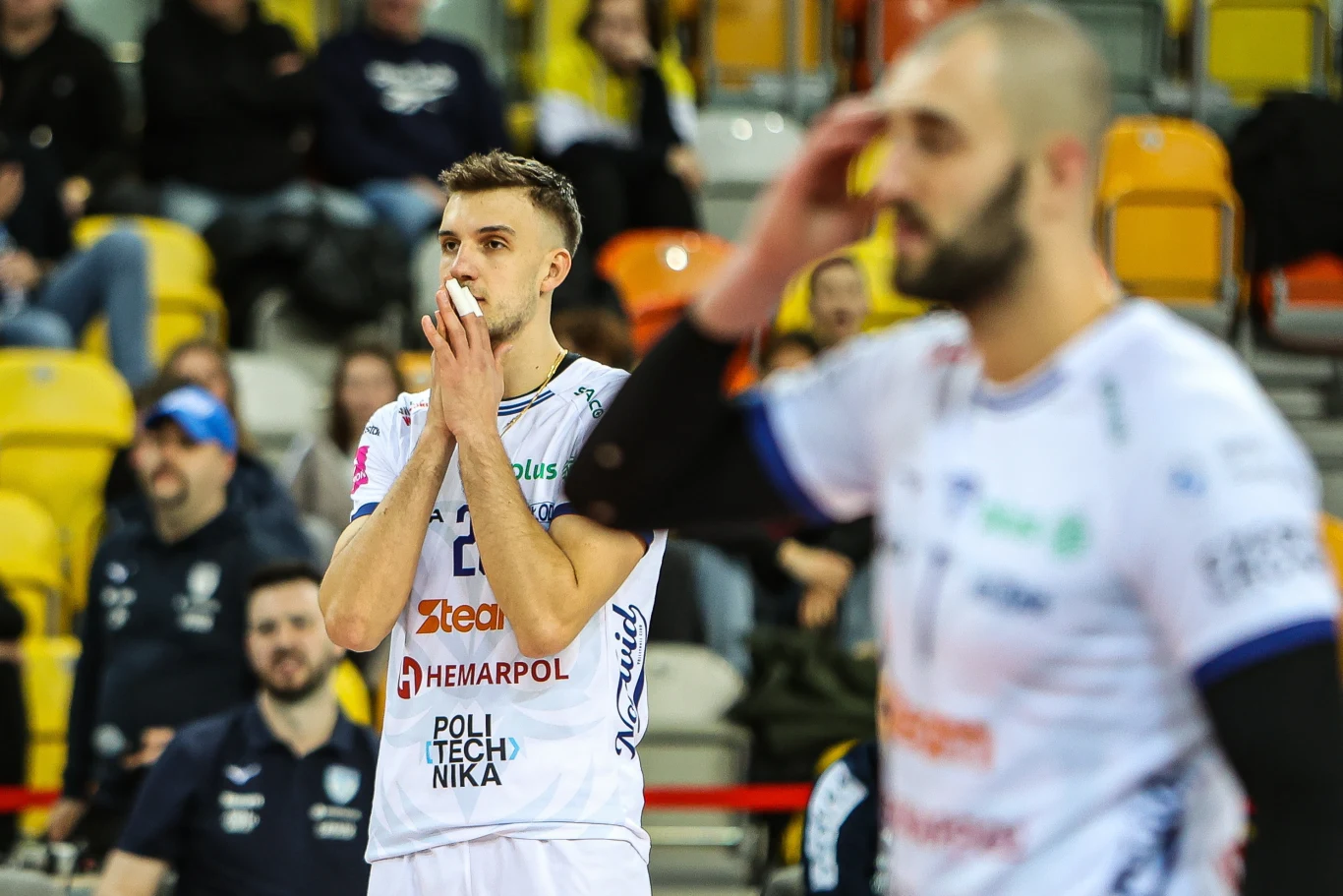 Patrik Indra został bohaterem Steam Hermapol Politechniki Częstochowa Siatkarz w białym stroju z opatrunkiem na nosie stoi na boisku, obok niego inny zawodnik, a w tle widoczna jest publiczność na trybunach.