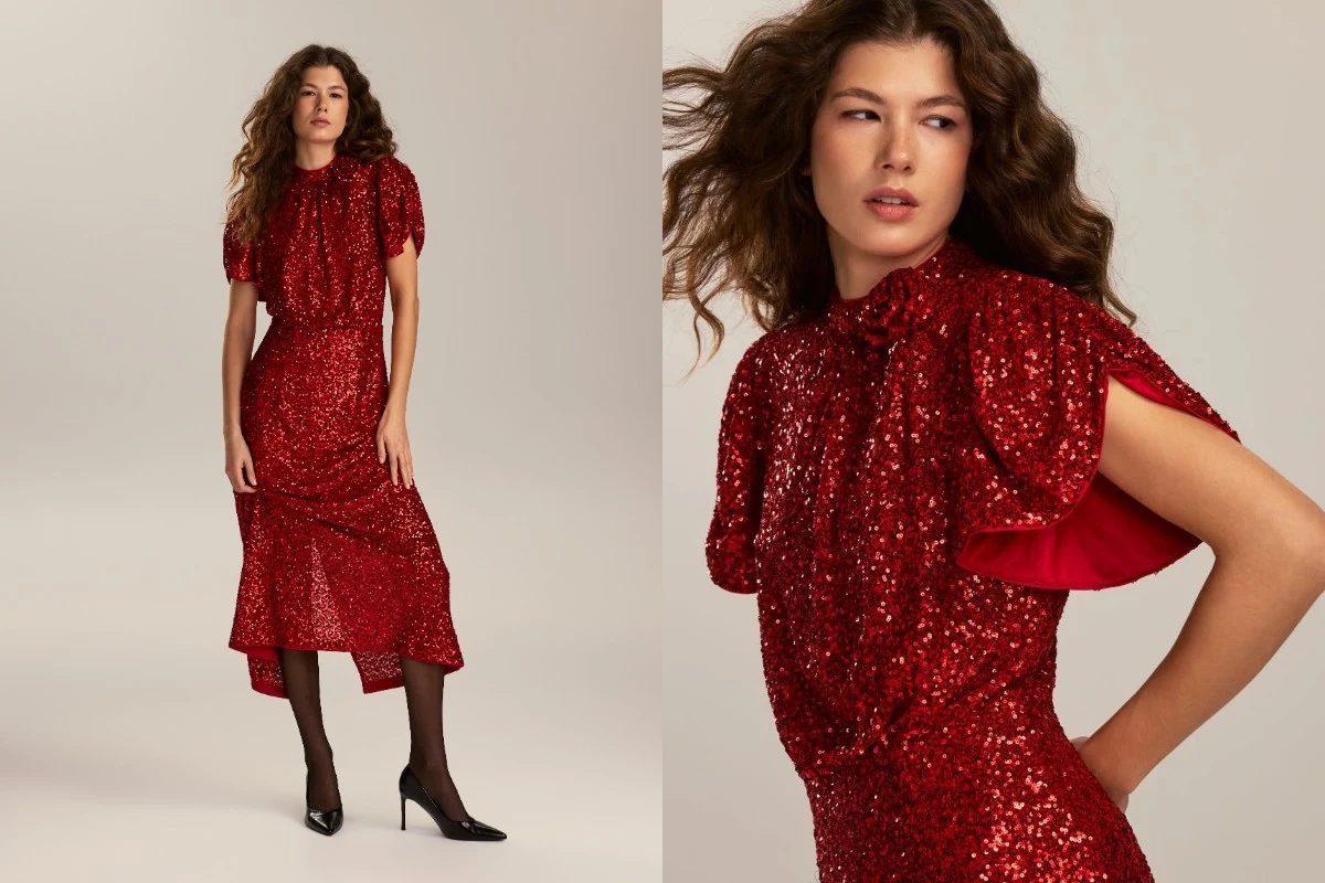 Kobieta z długimi, falowanymi włosami nosi cekinową czerwoną sukienkę o długości za kolano z krótkim rękawem i kokardą pod szyją, do zestawu założone czarne, kryjące rajstopy i czarne szpilki, tło jednolite, jasne.