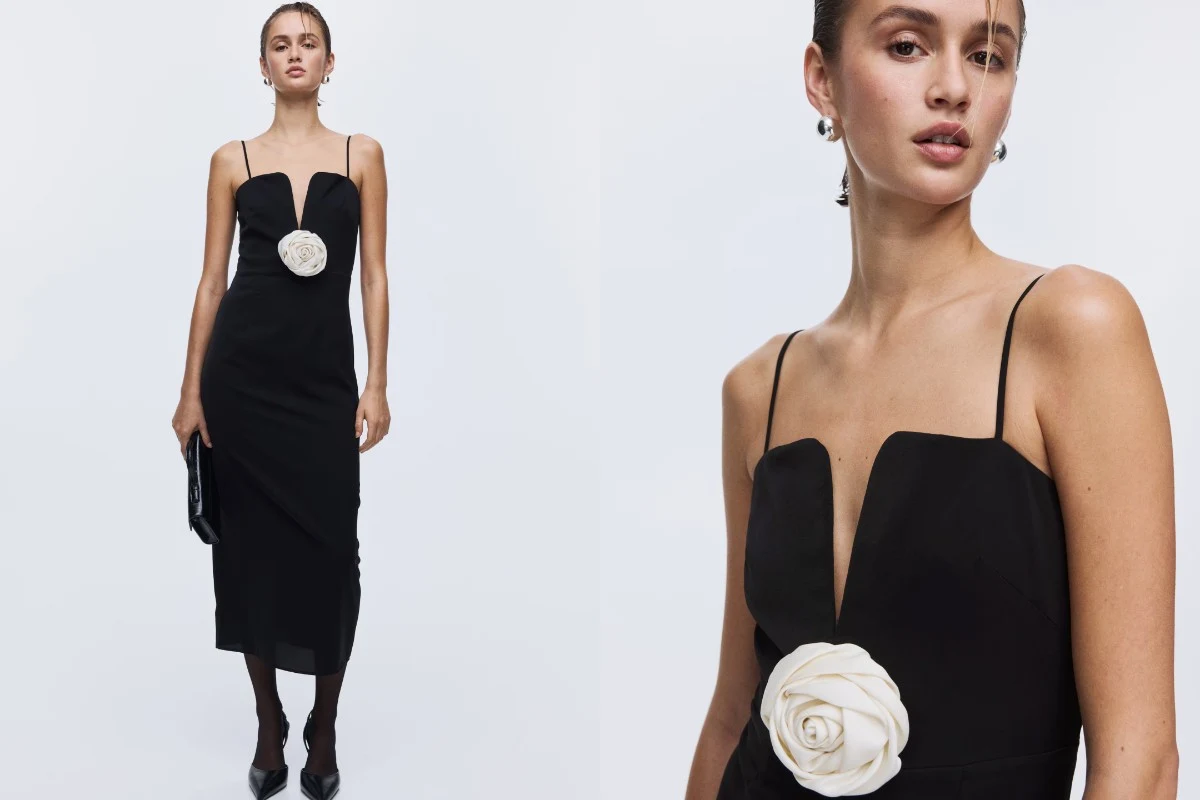 Elegancka kobieta ubrana w czarną sukienkę na cienkich ramiączkach, ozdobioną dużą białą różą przy dekolcie, trzyma w dłoni czarną kopertówkę. Stylizację uzupełniają subtelne kolczyki i klasyczne czarne szpilki.