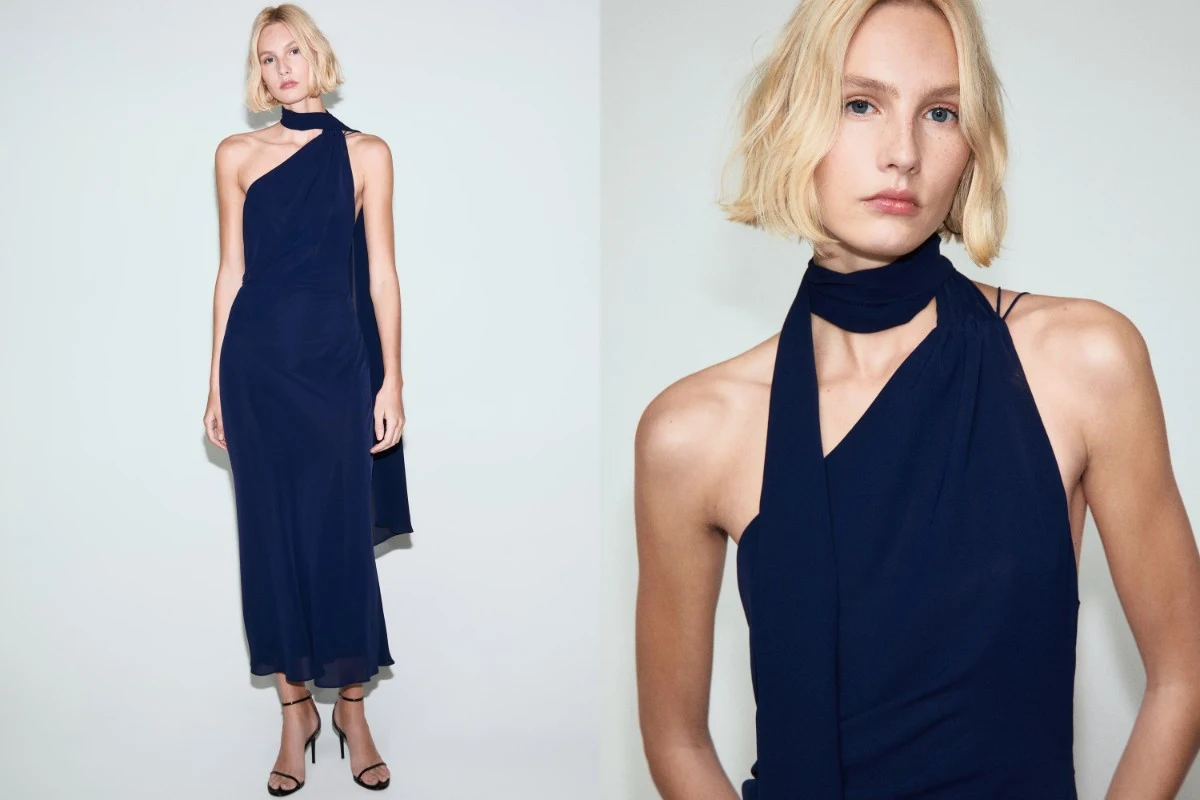 Młoda kobieta z jasnymi włosami nosząca długą, granatową suknię bez rękawów ze stylowym szalem oplatającym szyję, stojąca na tle jasnej ściany. Minimalistyczna stylizacja, elegancki i wyrafinowany wygląd, subtelne czarne sandały na obcasie.