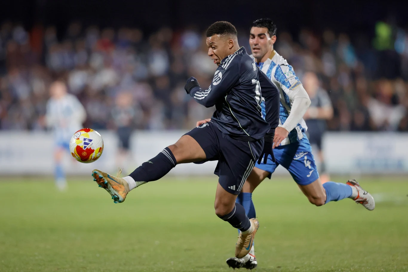 Piłkarz w granatowym stroju kopie piłkę, a za nim biegnie zawodnik przeciwnej drużyny w niebiesko-białym stroju. Akcja toczy się na boisku podczas meczu, w tle widoczna rozmyta publiczność.