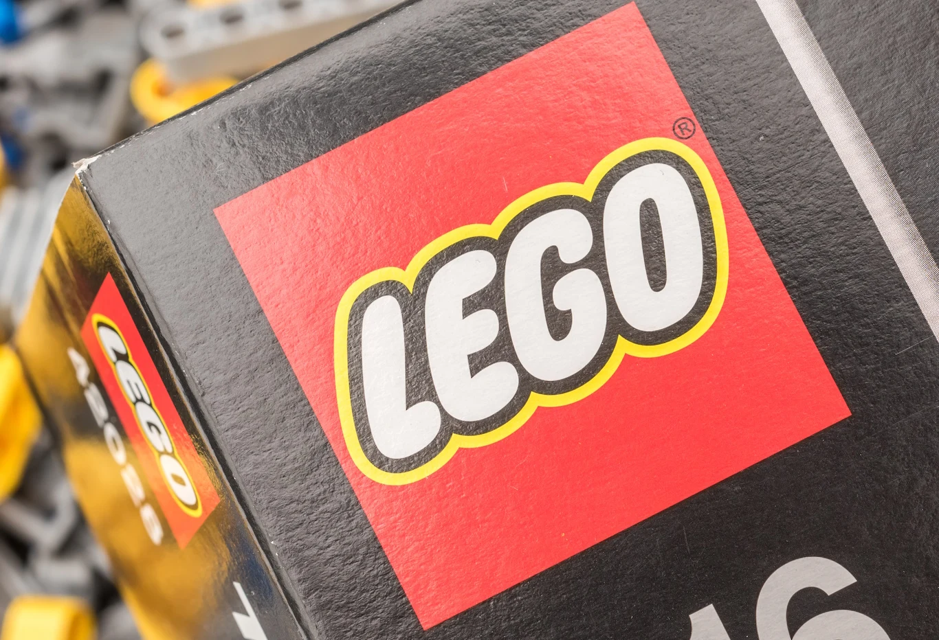 LEGO Logo LEGO na pudełku zestawu klocków, tło stanowią części zabawek w kolorach szarym, żółtym i niebieskim.