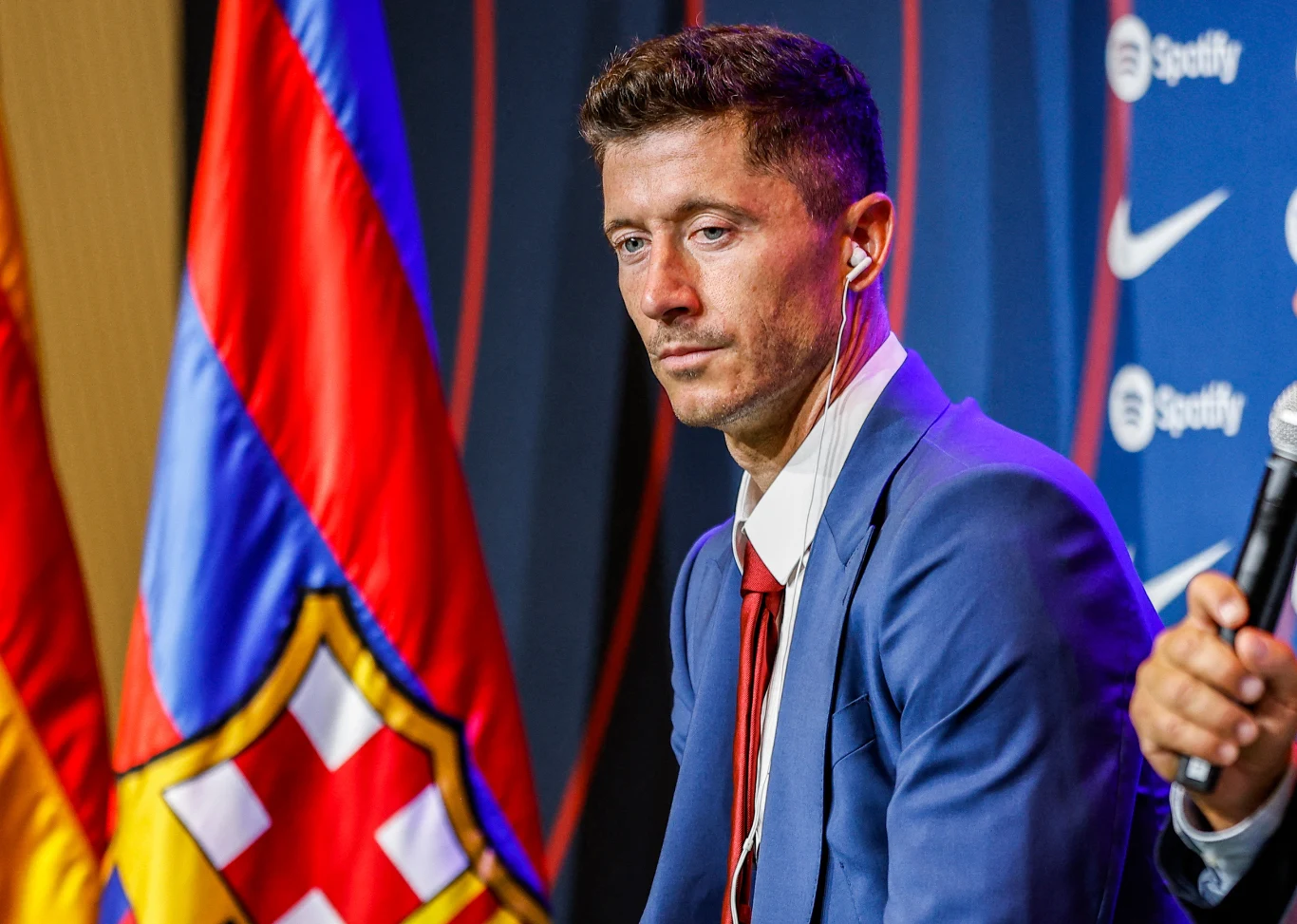 Mężczyzna w niebieskim garniturze i białej koszuli z czerwonym krawatem siedzi na tle flag FC Barcelony oraz ściany z logotypami sponsora i klubu, mając założone słuchawki.