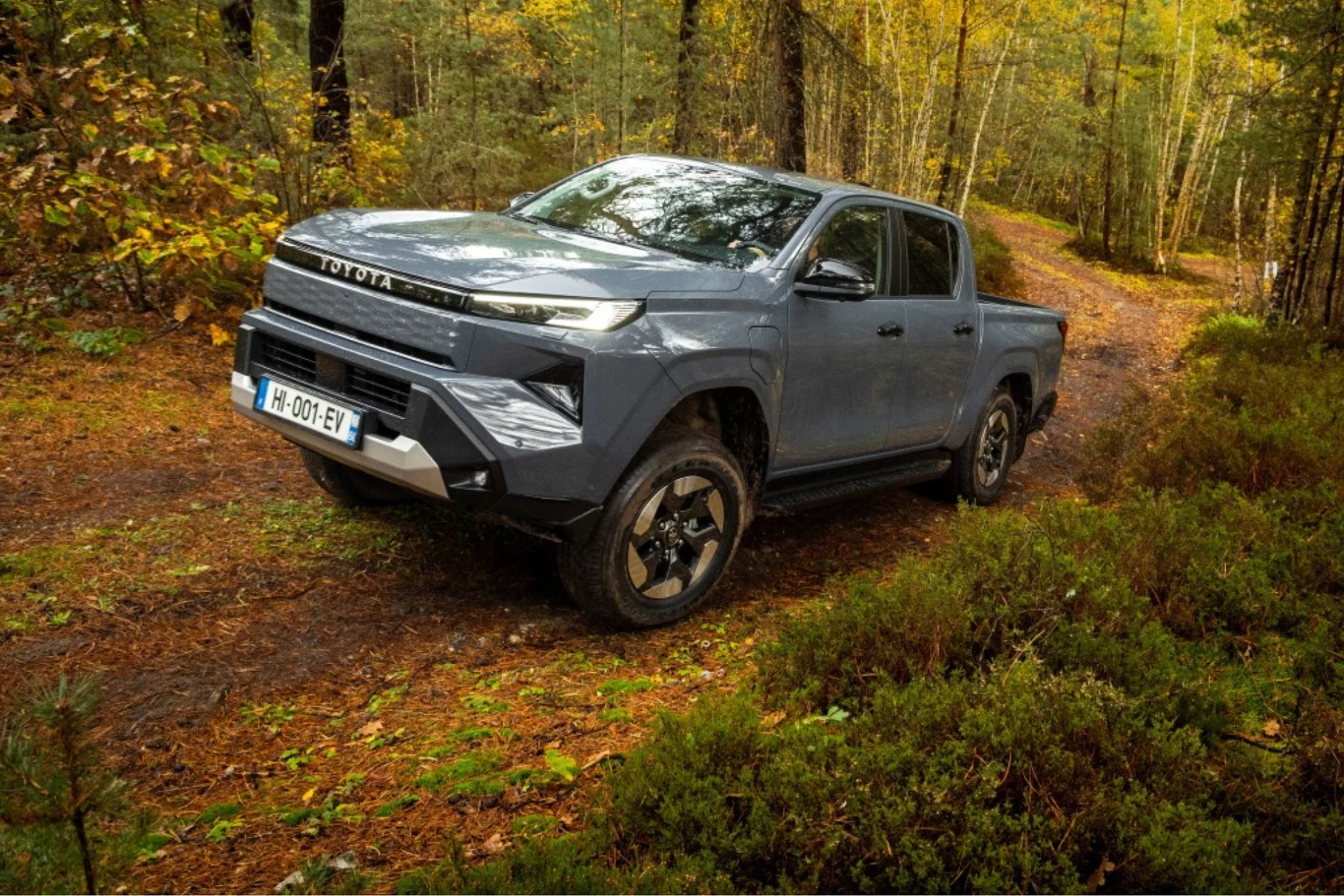 Elektryczna Toyota Hilux debiutuje w Polsce. Szary pickup Dacia z nowoczesnym designem stoi na leśnej, nieutwardzonej drodze otoczonej jesienną roślinnością i drzewami, auto wygląda solidnie i jest przeznaczone do jazdy terenowej.