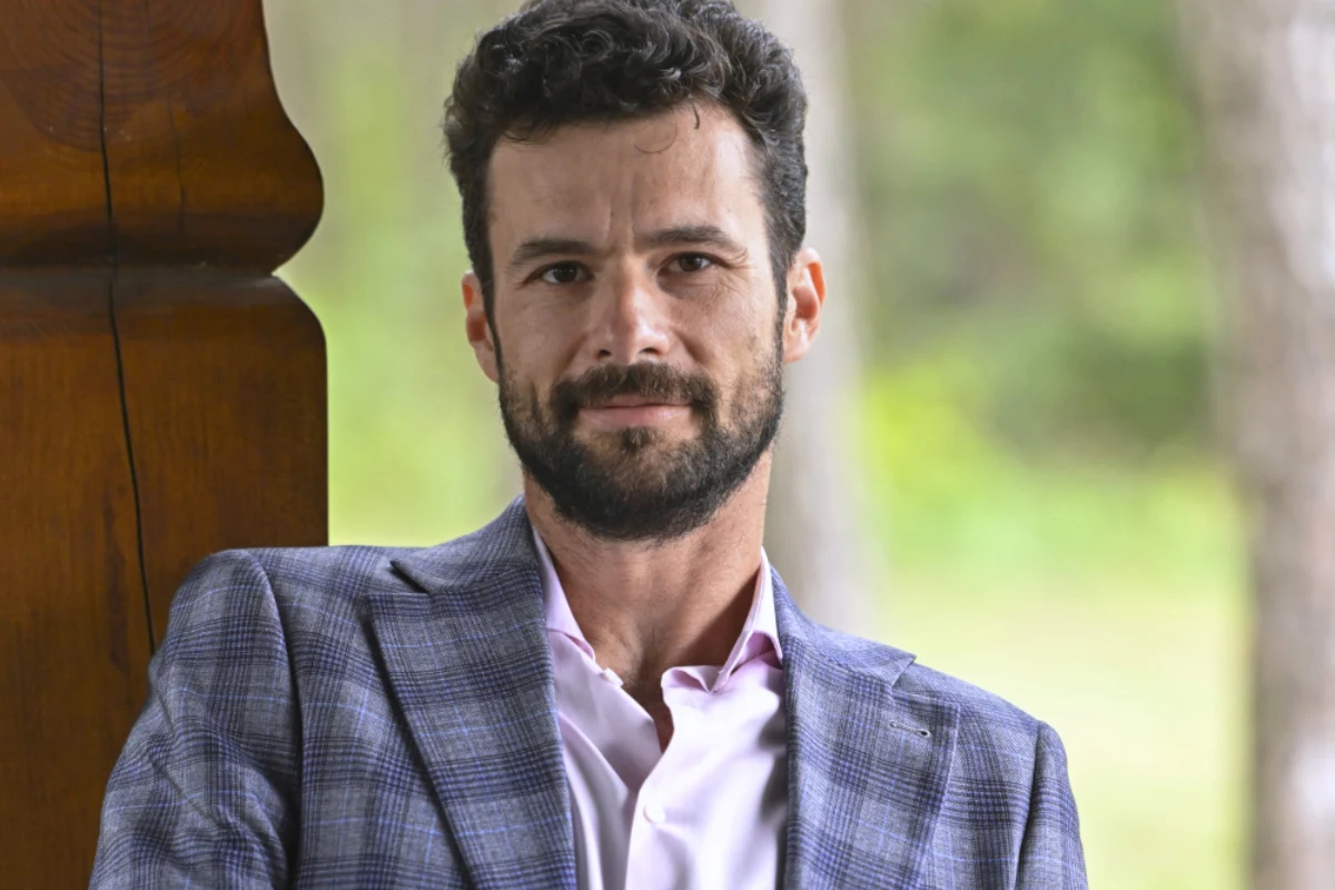 Roland z "Rolnik szuka żony" 12 Mężczyzna z brodą i kręconymi włosami w marynarce w kratę oraz jasnoróżowej koszuli, siedzący na tle rozmytego zielonego krajobrazu. Lekko się uśmiecha, patrząc wprost do obiektywu.