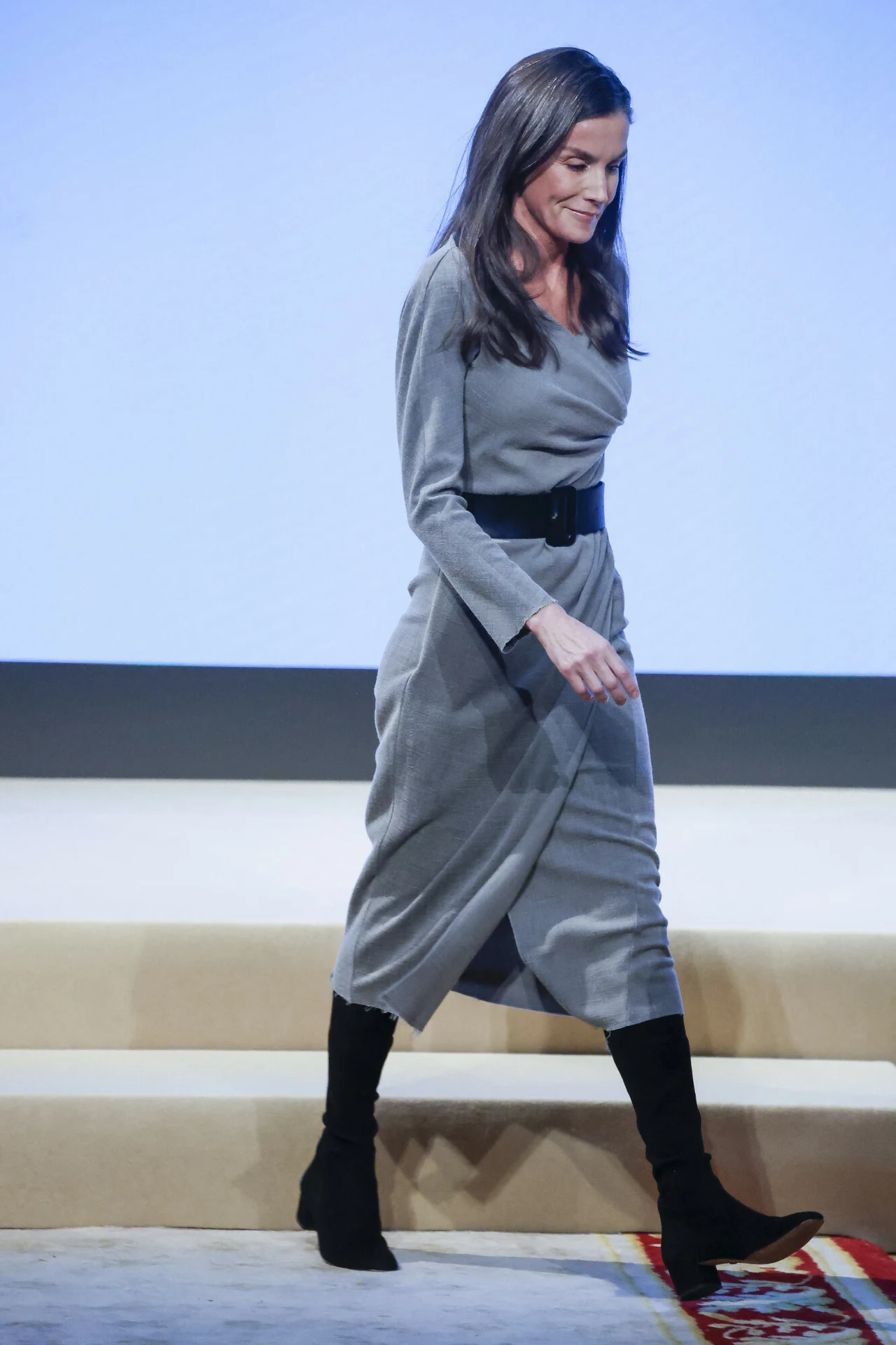 Kobieta w szarej sukience z czarnym paskiem i długimi czarnymi butami idzie po jasnej scenie w kierunku boku kadru. W tle jednolite, jasne tło, podłoże pokryte jasnym dywanem z czerwonym wzorem w jednym z rogów.