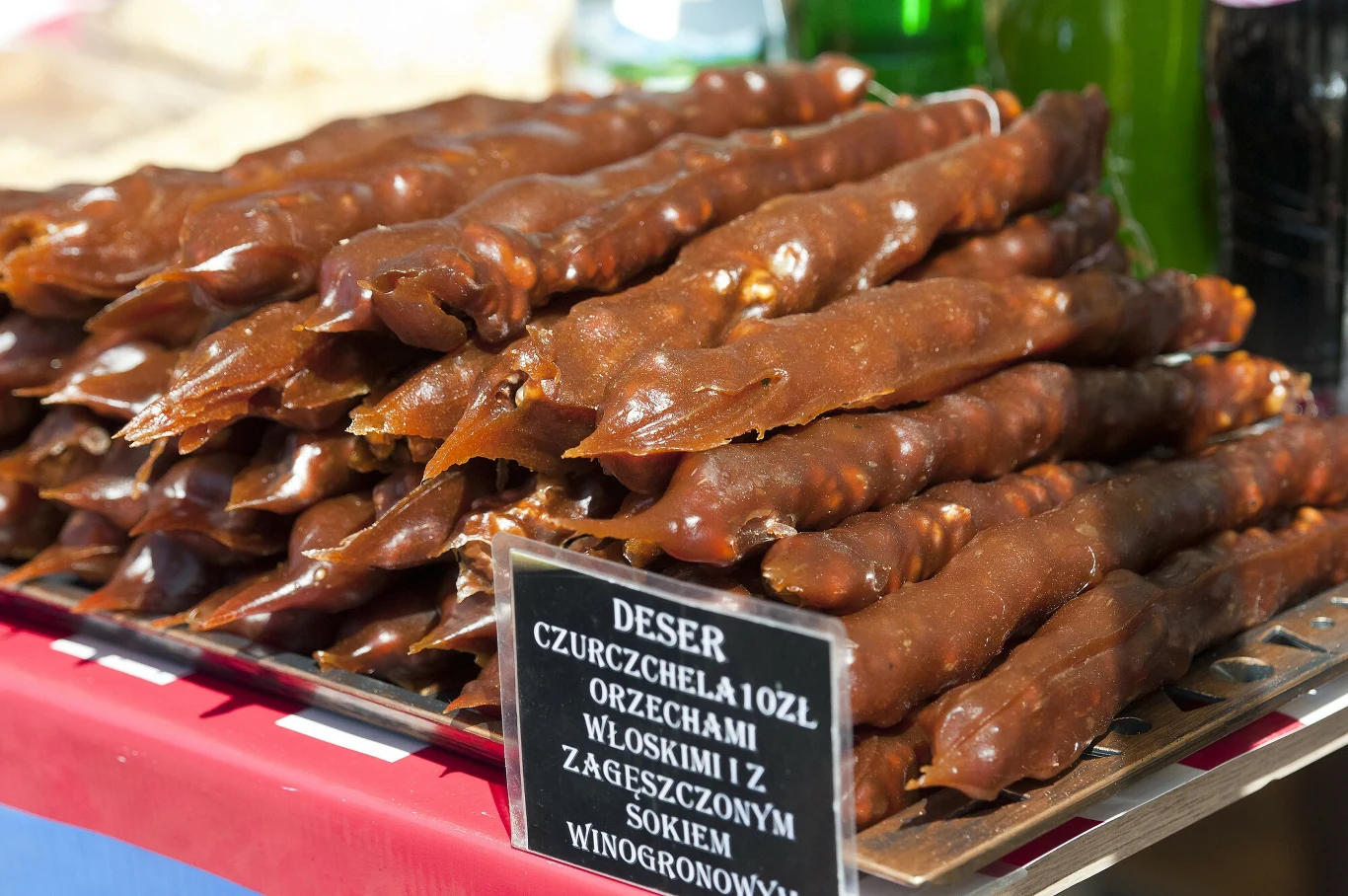 Stos deserów czurczchela z orzechami włoskimi pokrytych gęstym sosem z soku winogronowego, ustawiony na metalowej tacy, z widoczną etykietą informacyjną z nazwą i składnikami produktu.