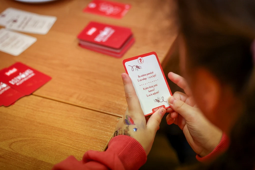 Inka i TPD tworzą przestrzeń do rozmów międzypokoleniowych Osoba w czerwonej bluzie trzyma w dłoni kartę z tekstem, siedząc przy drewnianym stole, na którym leżą pozostałe karty do gry.