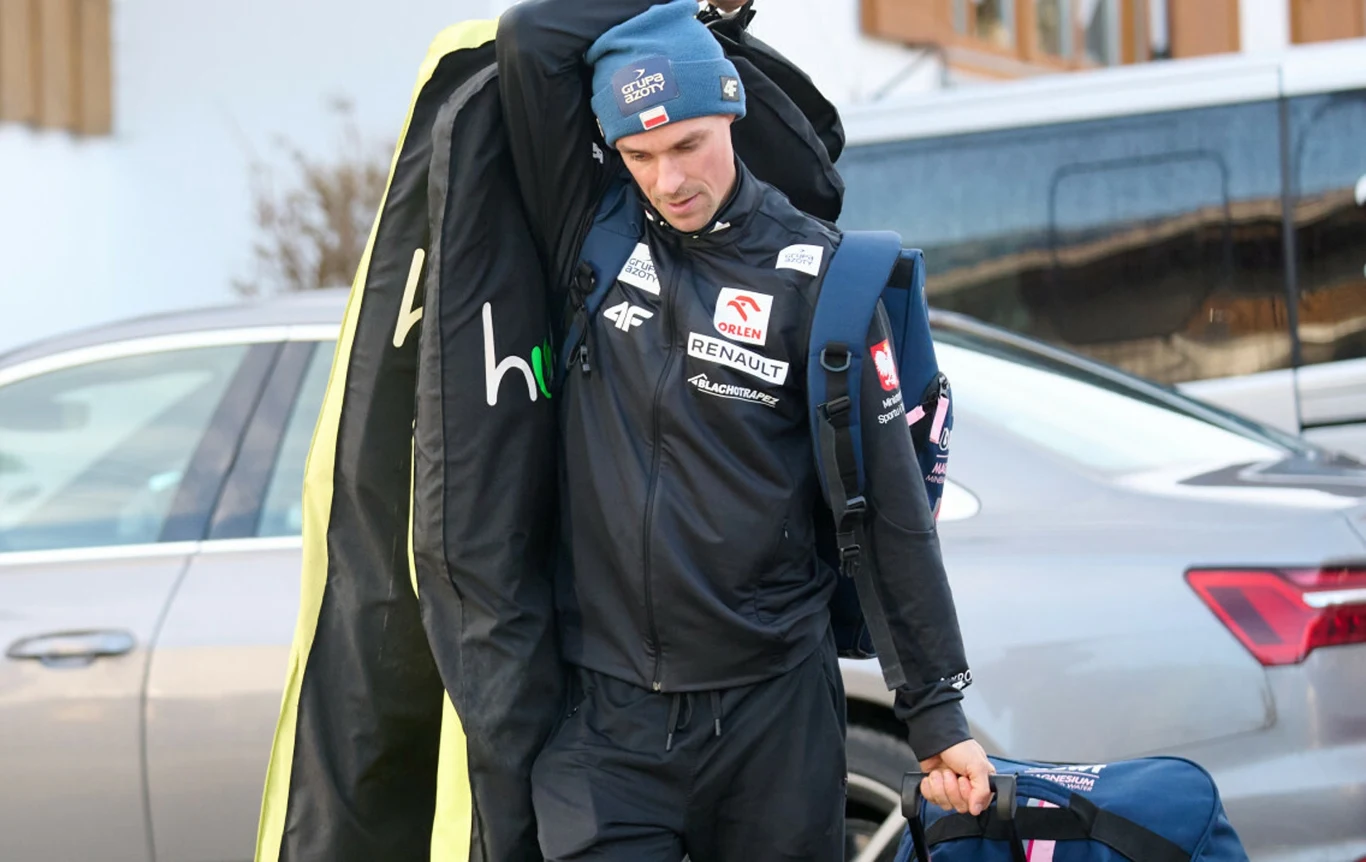 Mężczyzna w sportowej odzieży, z czapką na głowie niesie pokrowiec na sprzęt oraz torbę sportową, w tle zaparkowany samochód i budynki.