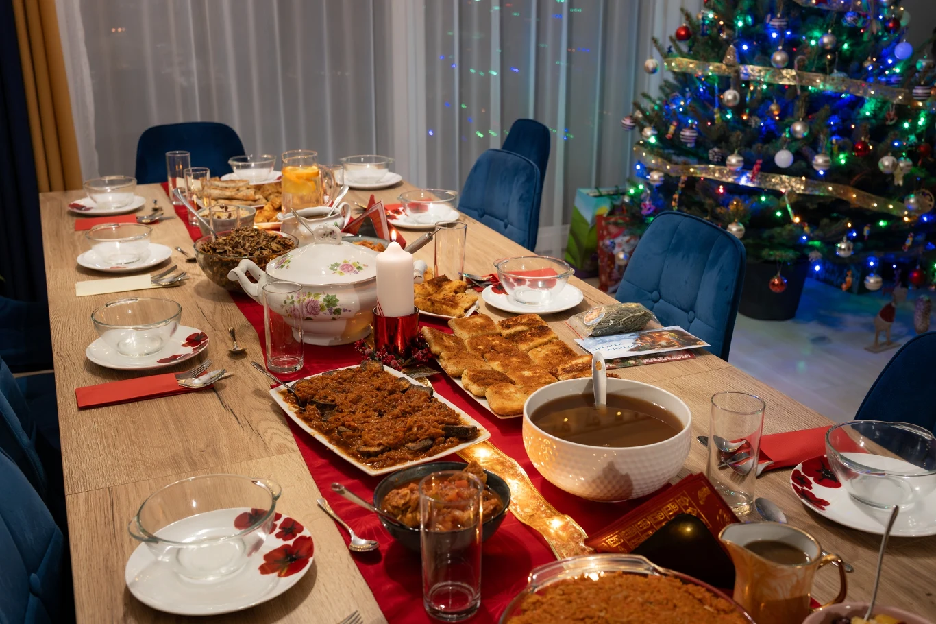 Świąteczny stół może stać się źródłem strat. Marnowanie żywności to poważny problem. Długi stół przykryty czerwonym bieżnikiem, starannie nakryty zastawą na kilka osób, wokół ustawione niebieskie krzesła. Po środku stołu znajdują się tradycyjne potrawy wigilijne, napoje oraz półmiski i wazony z jedzeniem. W tle oświetlona choinka ozdob...