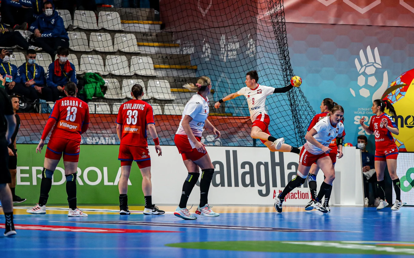 Akcja podczas meczu piłki ręcznej kobiet, zawodniczka w białym stroju unosi się w powietrzu w trakcie oddawania rzutu na bramkę, zawodniczki drużyny przeciwnej w czerwonych strojach obserwują sytuację na boisku, w tle trybuny i kibice.