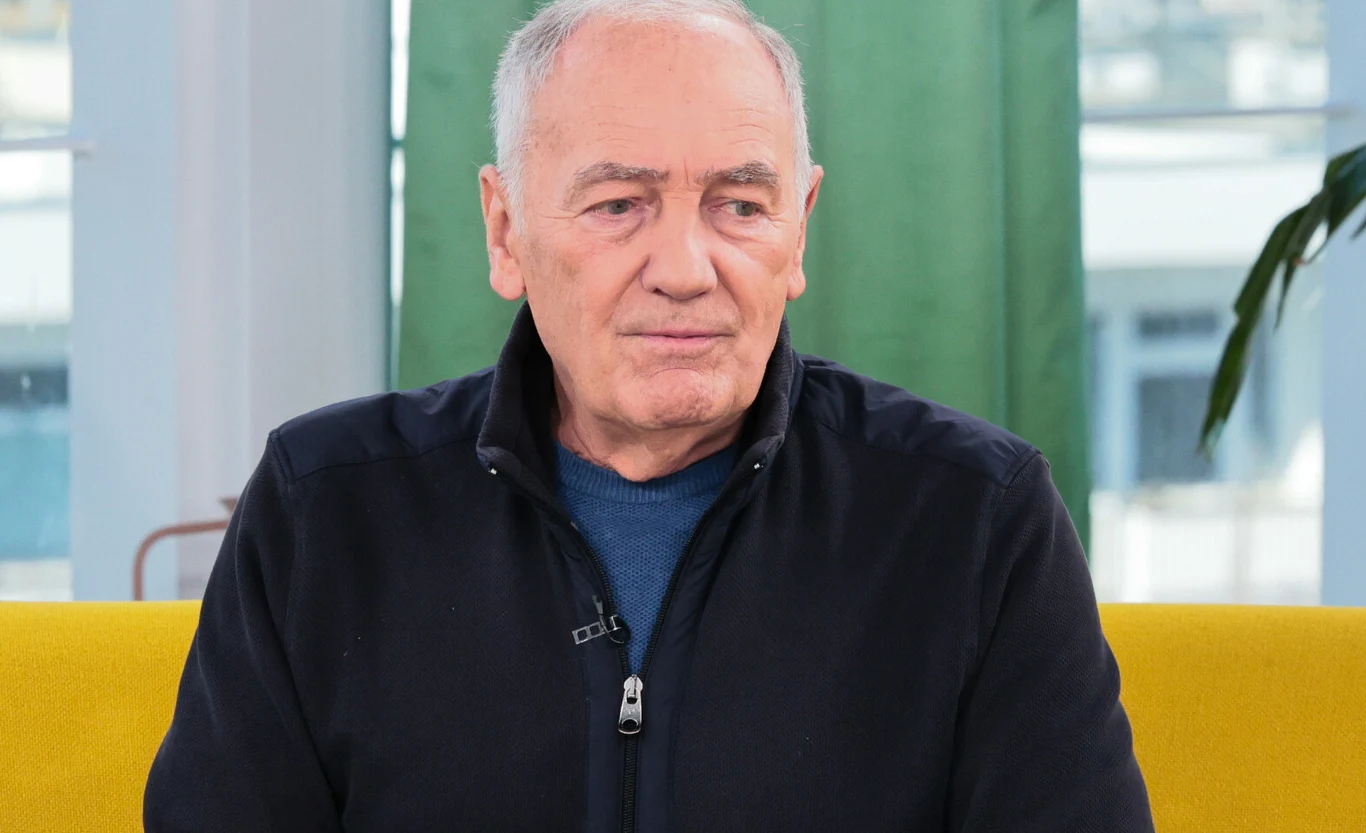 Karol Strasburger Starszy mężczyzna siedzący na żółtej sofie, ubrany w ciemną bluzę i niebieską koszulkę, w tle zielona zasłona i roślina doniczkowa.