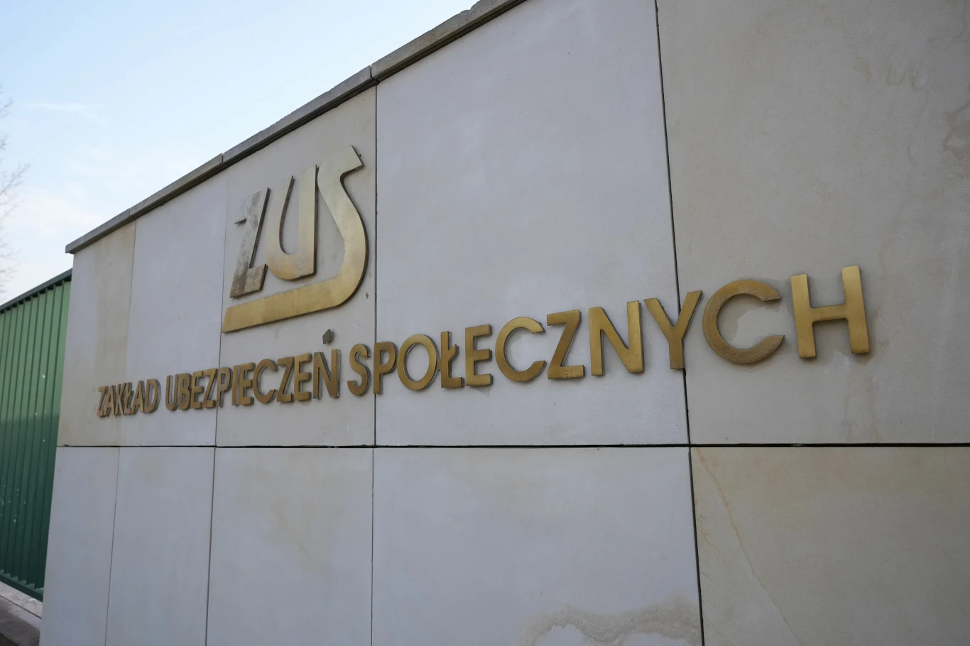 ZUS: FUS pozostaje w stabilnej kondycji (zdj. ilustracyjne) Złoty napis Zakład Ubezpieczeń Społecznych umieszczony na jasnej ścianie budynku, obok zielonego metalowego ogrodzenia.