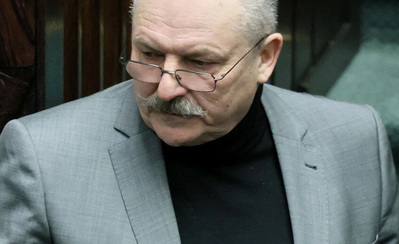 Marek Jakubiak Starszy mężczyzna z siwymi włosami i wąsem, ubrany w szary garnitur oraz czarny golf, ma na nosie okulary, a jego wyraz twarzy jest skupiony.