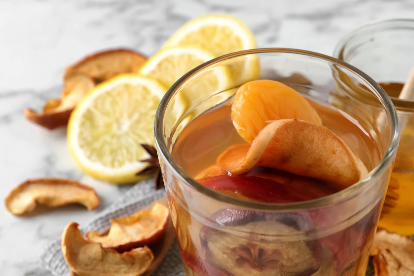 Szklanka kompotu z suszonych owoców, w której znajdują się kawałki suszonych jabłek, moreli i śliwek, na drugim planie widoczne plasterki cytryny oraz kawałki suszonych owoców na szarej serwetce.