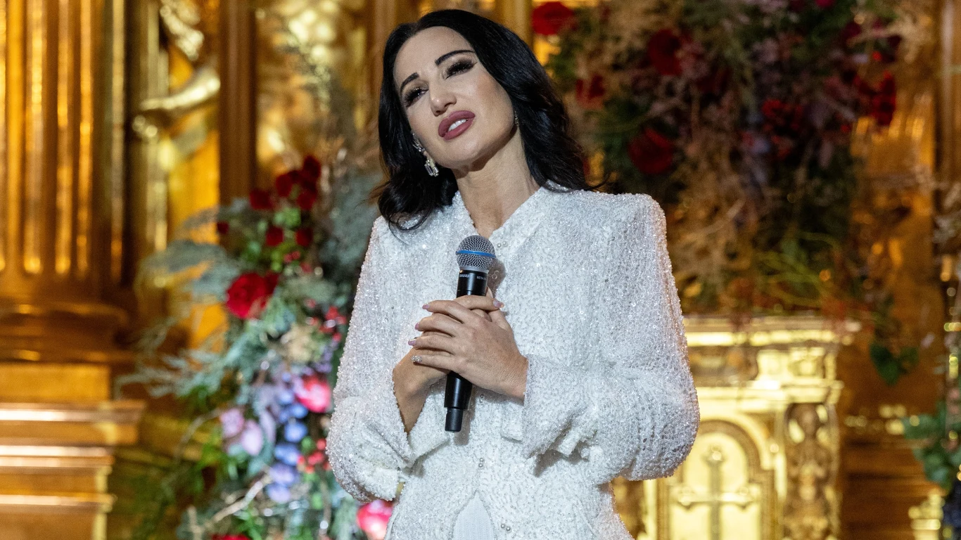 Justyna Steczkowska Kobieta w białym, eleganckim stroju trzyma mikrofon, stoi na tle bogato zdobionego, złotego wnętrza z kwiatowymi kompozycjami, sprawiając wrażenie śpiewającej lub przemawiającej.