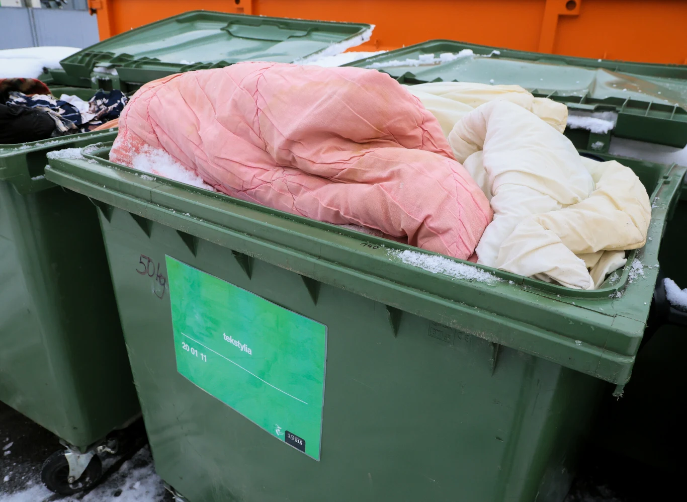 Gminy mają ustawowy obowiązek segregacji odpadów tekstylnych i odzieży Zielony plastikowy kontener na śmieci stojący na zewnątrz, na którego wierzchu leżą porzucone kołdry w jasnych kolorach, na tle śniegu i innych pojemników.