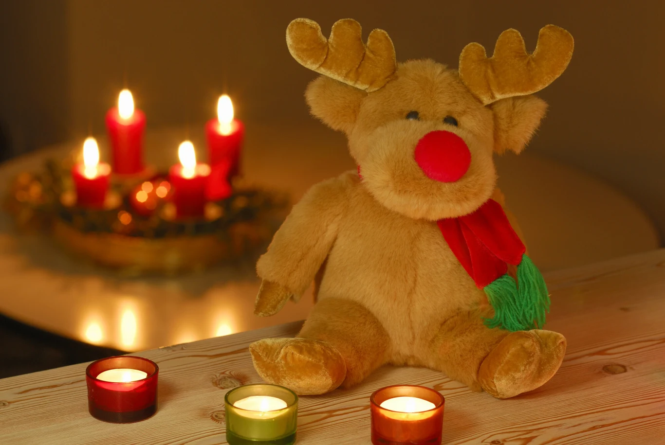 Pluszowy renifer z czerwoną chustką na szyi siedzi na drewnianym stole, przed nim stoją trzy zapalone świeczki w kolorowych szklankach, w tle świecą się czerwone świece ustawione na ozdobnej tacy.