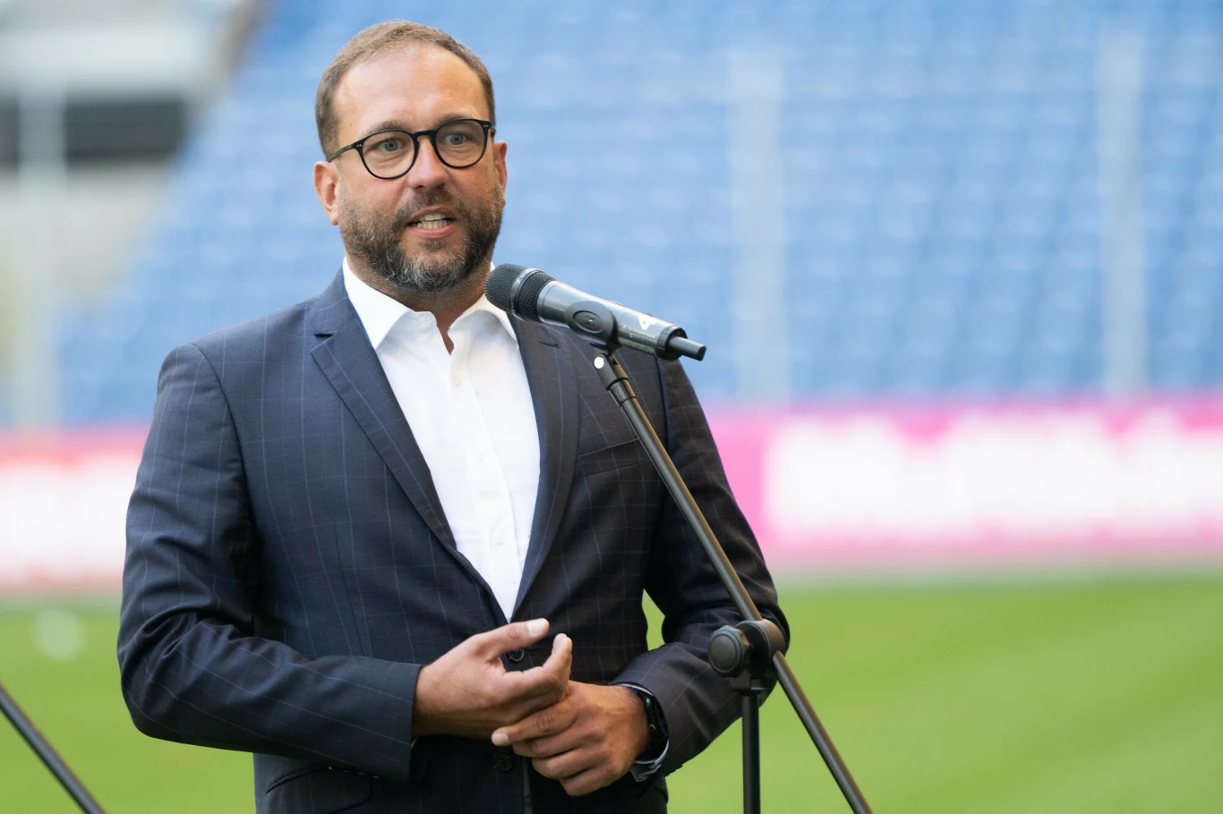 Mężczyzna w ciemnym garniturze i okularach przemawia do mikrofonu, tło stanowią trybuny stadionu
