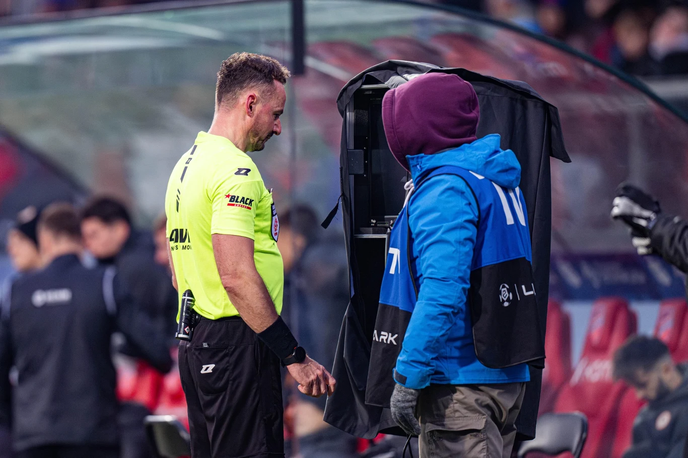 Sędzia piłkarski w żółtej koszulce obserwuje monitor VAR przy linii bocznej boiska, obok stoi operator w niebieskiej kamizelce i kurtce z kapturem, w tle widoczne są rozmazane sylwetki innych osób oraz stadionowe ławki.
