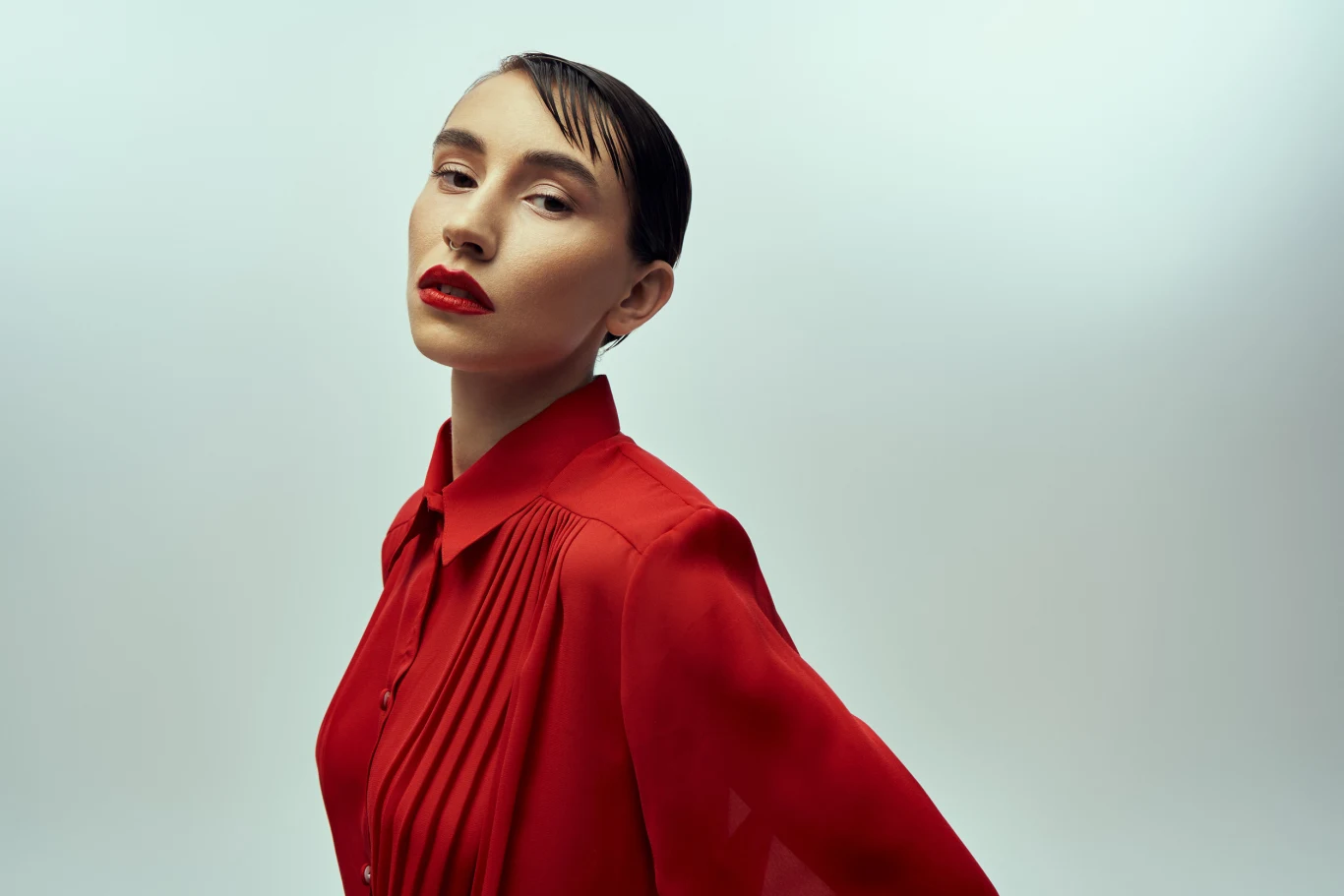 Kobieta z ciemnymi, zaczesanymi do tyłu włosami pozuje w eleganckiej, czerwonej koszuli na jednolitym, jasnym tle. Jej wyraz twarzy jest poważny, podkreślony intensywną czerwoną szminką.