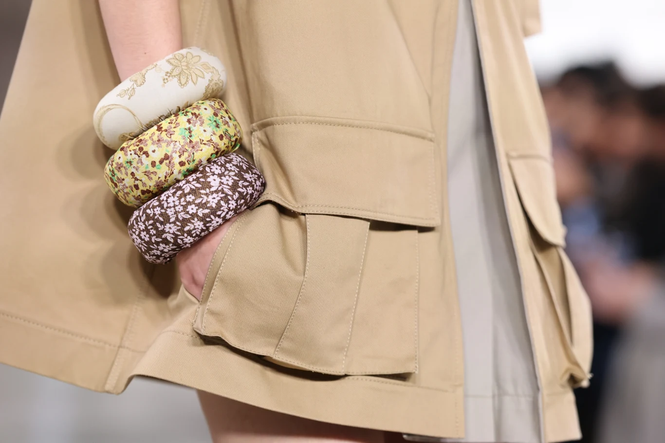 Ręka osoby nosząca kilka szerokich, materiałowych bransoletek w różnych wzorach kwiatowych, opierająca się na beżowym płaszczu z dużą kieszenią.