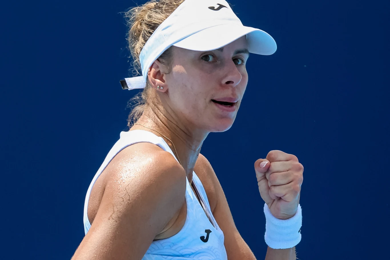 Magda Linette bierze udział w turnieju WTA 250 w Hobart Zbliżenie na tenisistkę ubranej w białą koszulkę i czapkę z daszkiem, wykonującą gest triumfu z zaciśniętą pięścią, na tle niebieskiego kortu.