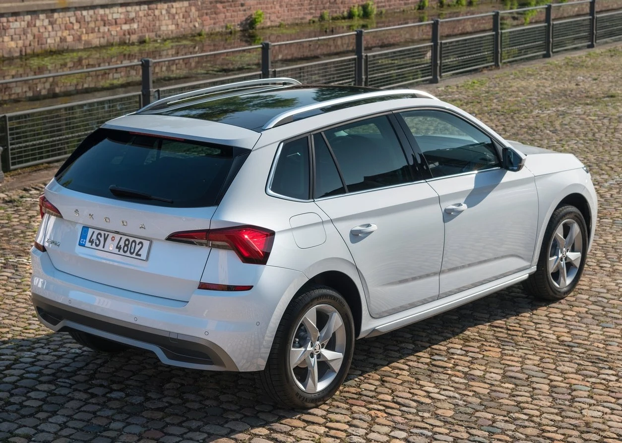 Białe auto typu SUV marki Škoda zaparkowane na brukowanej nawierzchni, widoczne z tyłu pod kątem, z zamkniętymi szybami i przyciemnionymi szybami bocznymi, w tle ogrodzenie i ceglana ściana.