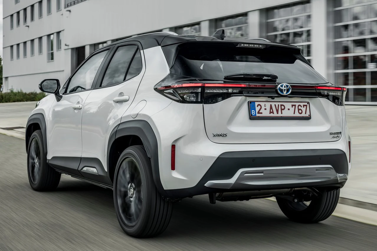 Biały samochód osobowy Toyota Yaris Cross na czarnych felgach porusza się po miejskiej drodze, widoczny od tyłu, w otoczeniu nowoczesnych budynków i przeszklonych fasad.