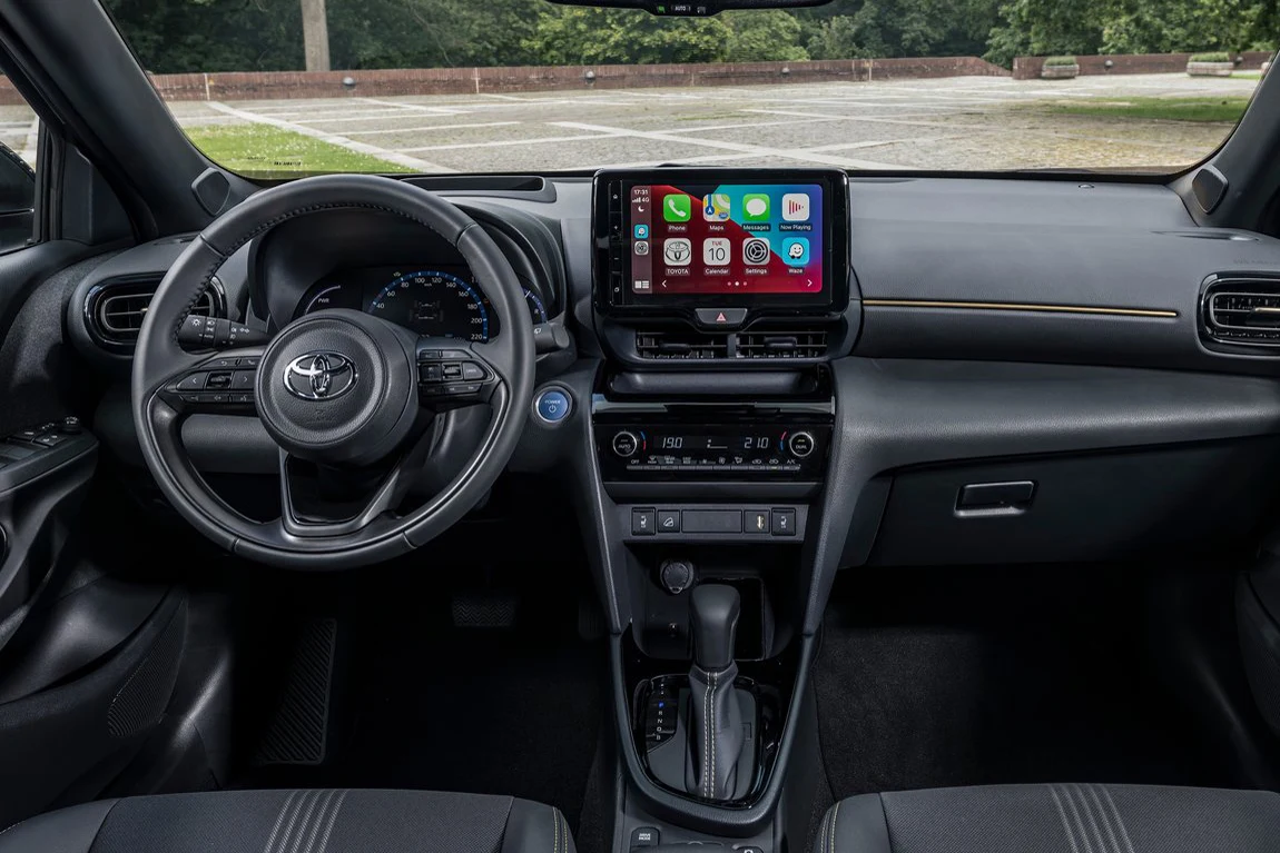 Wnętrze nowoczesnego samochodu marki Toyota z widoczną kierownicą, cyfrowym wyświetlaczem na desce rozdzielczej oraz dużym ekranem dotykowym systemu multimedialnego pośrodku konsoli. Fotele wykończone ciemnym materiałem z jasnymi przeszyciami, a w tle ...