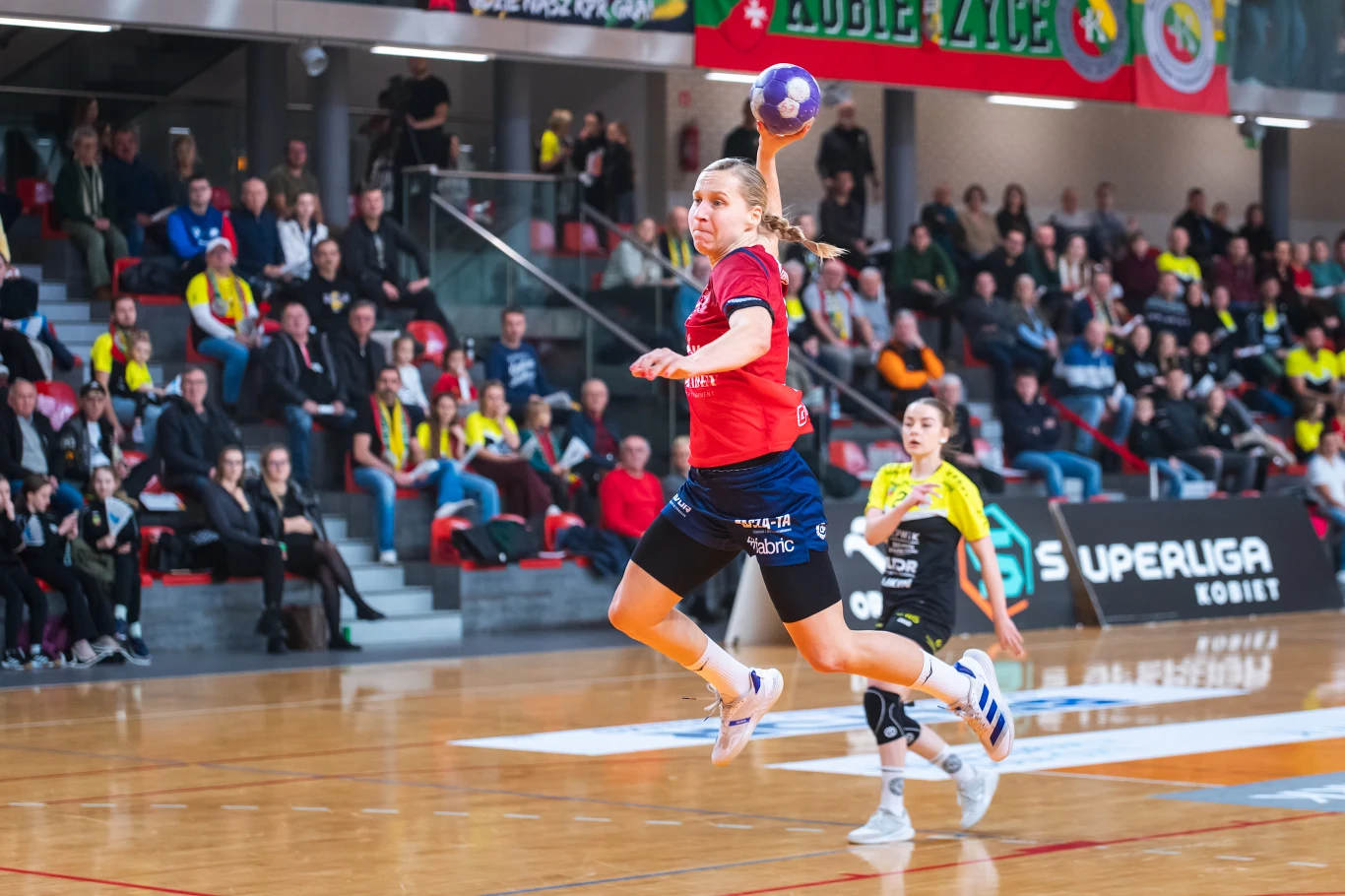 KPR Gminy Kobierzyce – MKS Piotrcovia Piotrków Tryb Zawodniczka piłki ręcznej w czerwonej koszulce wyskakuje przygotowując się do rzutu piłką, na tle widocznych trybun z kibicami i innymi zawodniczkami na boisku sportowym.