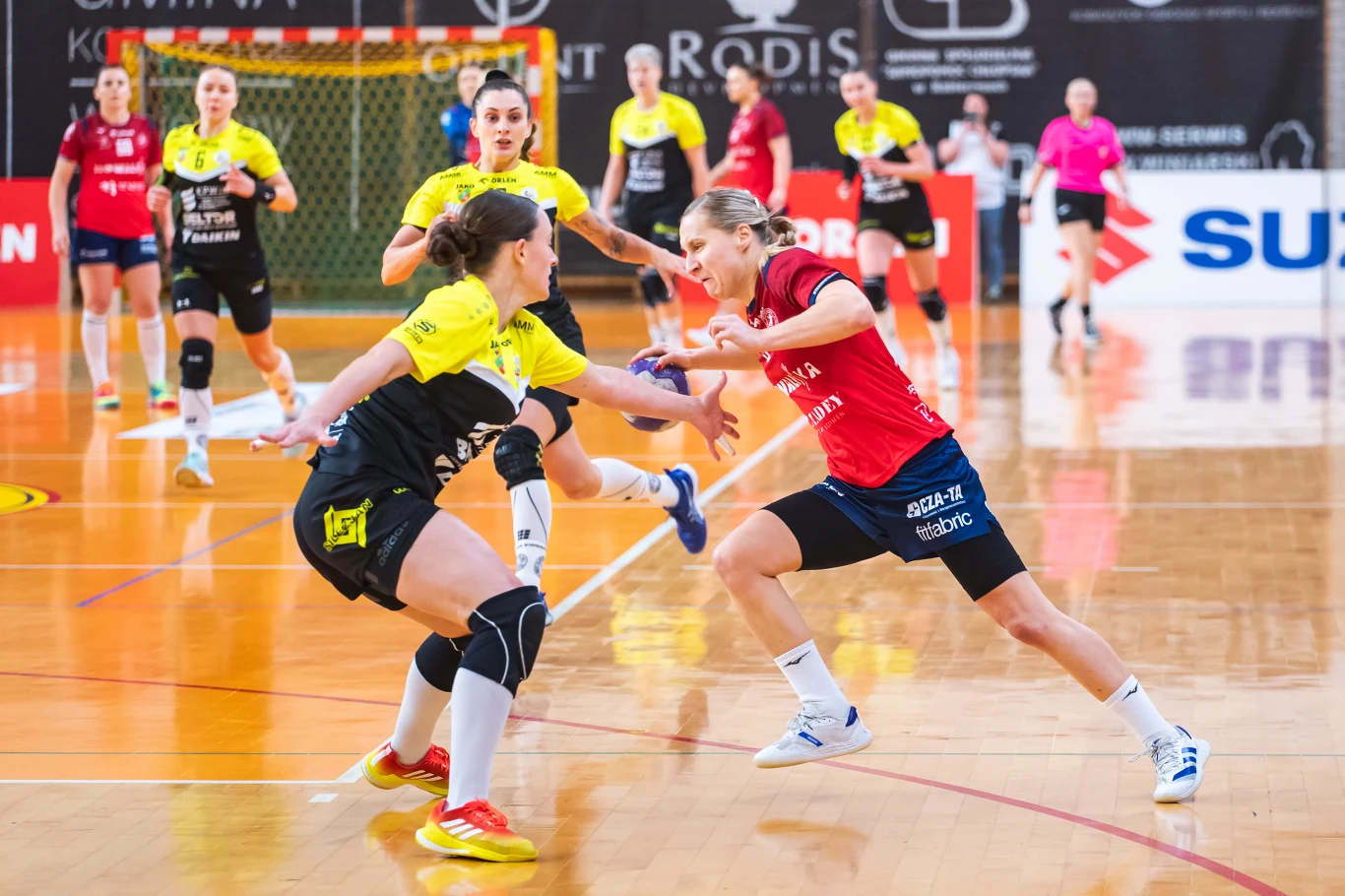 KPR Gminy Kobierzyce – MKS Piotrcovia Piotrków Tryb Intensywna akcja podczas meczu piłki ręcznej kobiet, dwie zawodniczki walczą o piłkę na parkiecie, pozostałe obserwują sytuację lub są gotowe do dalszej gry, w tle widoczna bramka, sędzia oraz reklamy sponsorów.