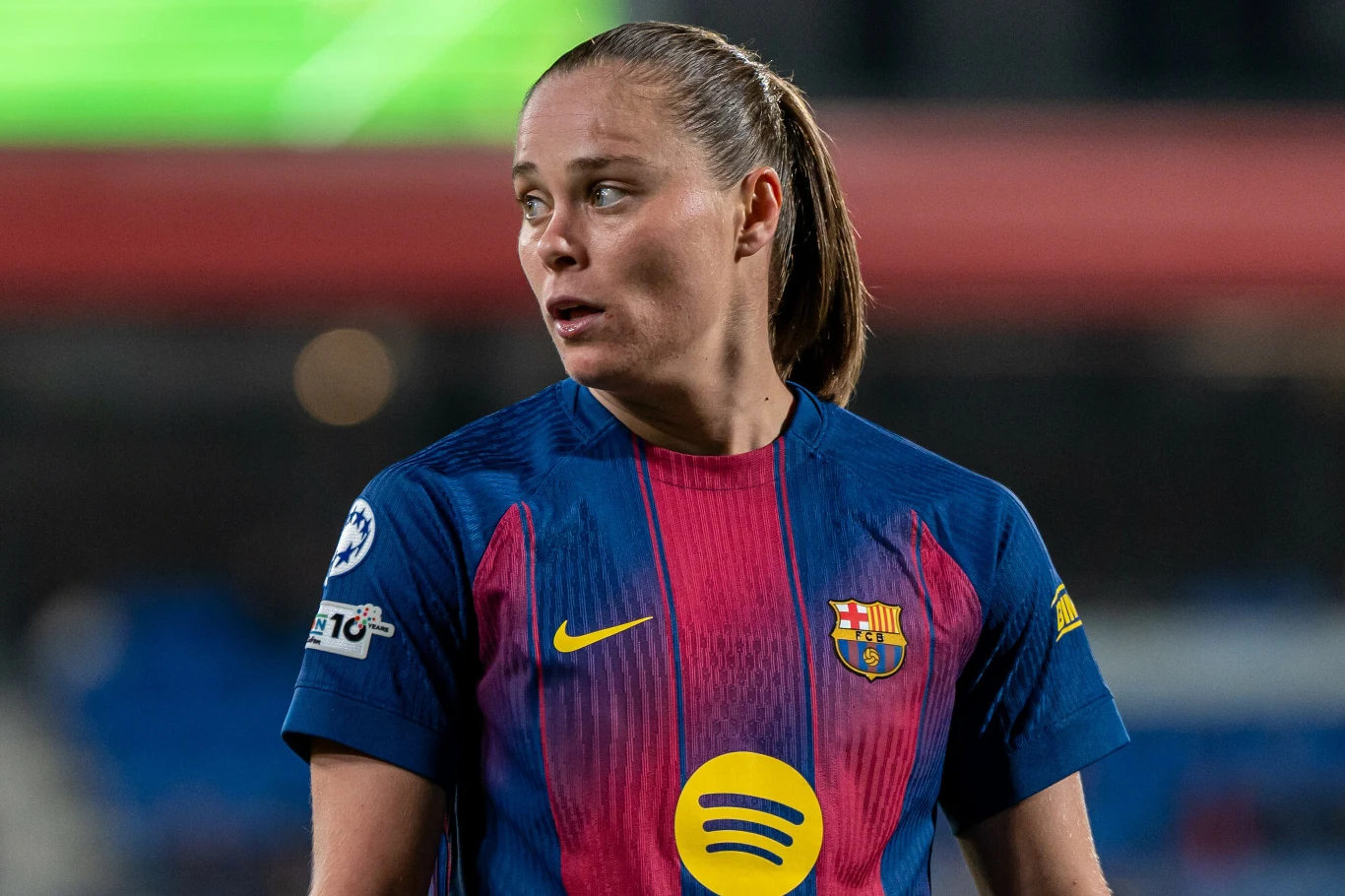 Piłkarka w niebiesko-czerwonej koszulce FC Barcelony z logo Spotify na klatce piersiowej stoi na boisku podczas meczu, skierowana bokiem, z poważnym wyrazem twarzy.