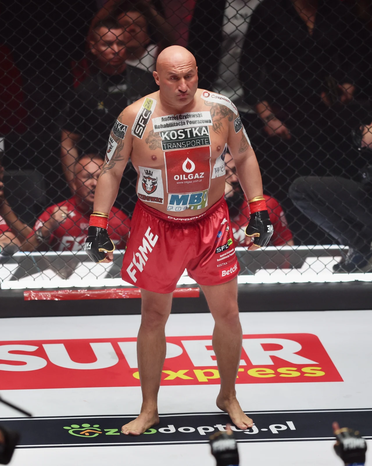 Łysy mężczyzna o atletycznej budowie ciała przygotowuje się do walki w klatce MMA, ubrany w czerwone spodenki z licznymi reklamami, jego ciało jest wytatuowane i pokryte logotypami sponsorów. W tle widać publiczność za siatką oktagonu.