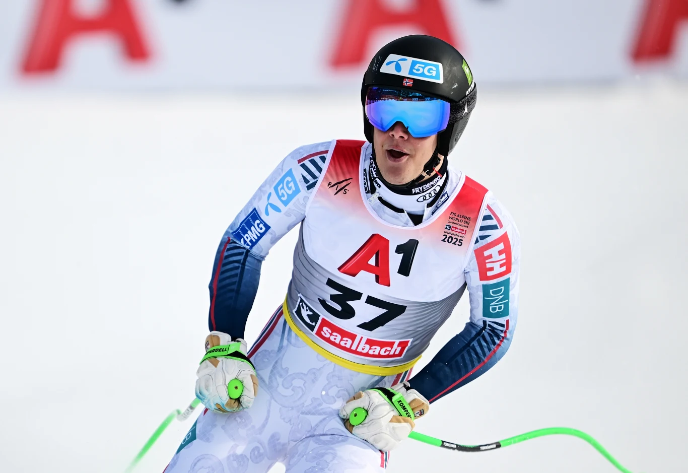 Narciarz w kasku i goglach podczas zawodów, ubrany w biały strój z numerem 37, trzyma kijki, wokół śnieżne tło i odblaskowe elementy stroju sportowego