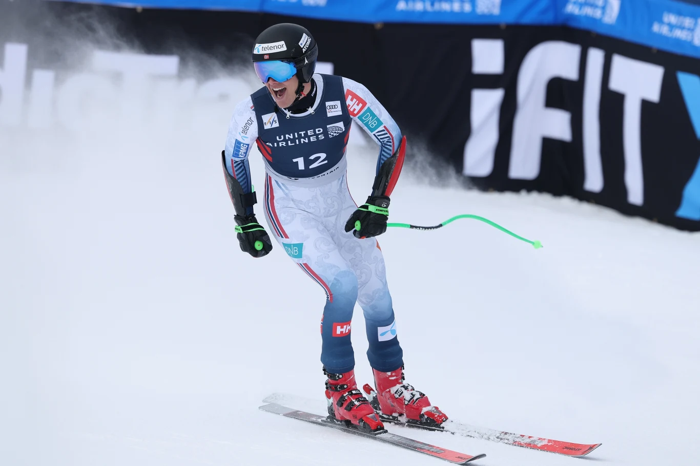 Narciarz alpejski w specjalnym kombinezonie oraz kasku, tuż po przekroczeniu linii mety podczas zawodów, trzymający kijki, z widocznym numerem startowym 12, dynamiczny ruch i intensywna mgła śnieżna w tle.