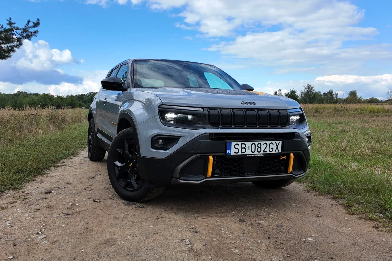 Samochód terenowy Jeep o szarej karoserii z czarnymi i żółtymi akcentami stoi na nieutwardzonej drodze, w tle widać pola i niską linię drzew pod niebieskim niebem z chmurami.