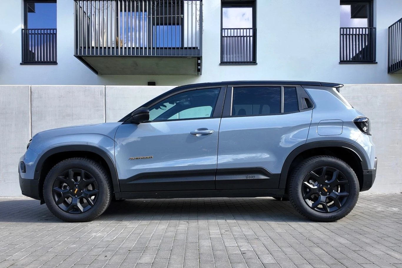 Srebrny SUV zaparkowany na brukowanym podjeździe przed nowoczesnym budynkiem z balkonami i białymi ścianami. Samochód widoczny z boku, z wyraźnym logo marki na drzwiach oraz ciemnymi felgami.