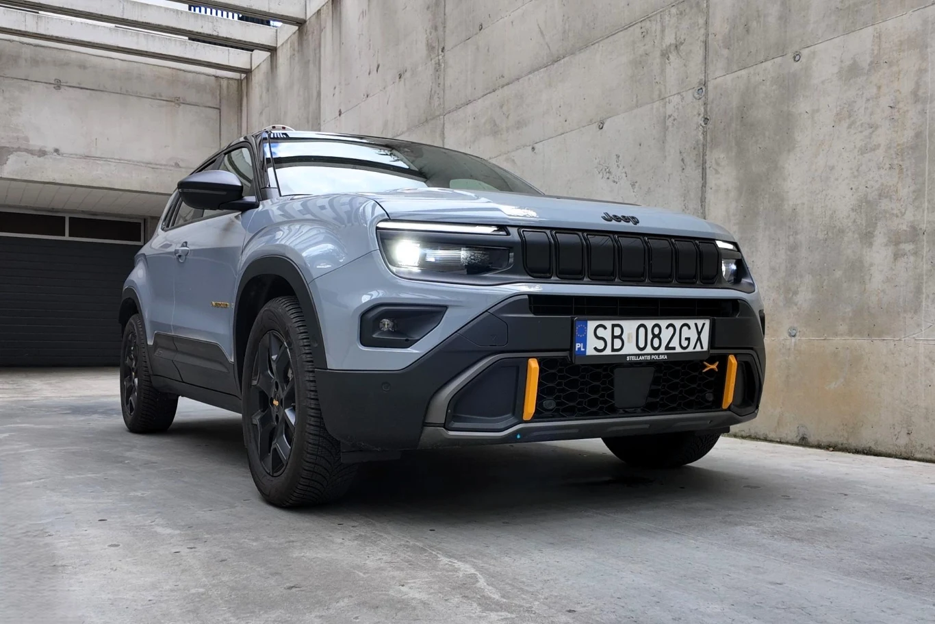 Nowoczesny samochód terenowy Jeep w jasnym kolorze, ustawiony na betonowym parkingu w otoczeniu surowych, betonowych ścian. Wyraźnie widoczne detale przodu, w tym charakterystyczna osłona chłodnicy i światła LED. Na przednim zderzaku żółte akcenty styl...
