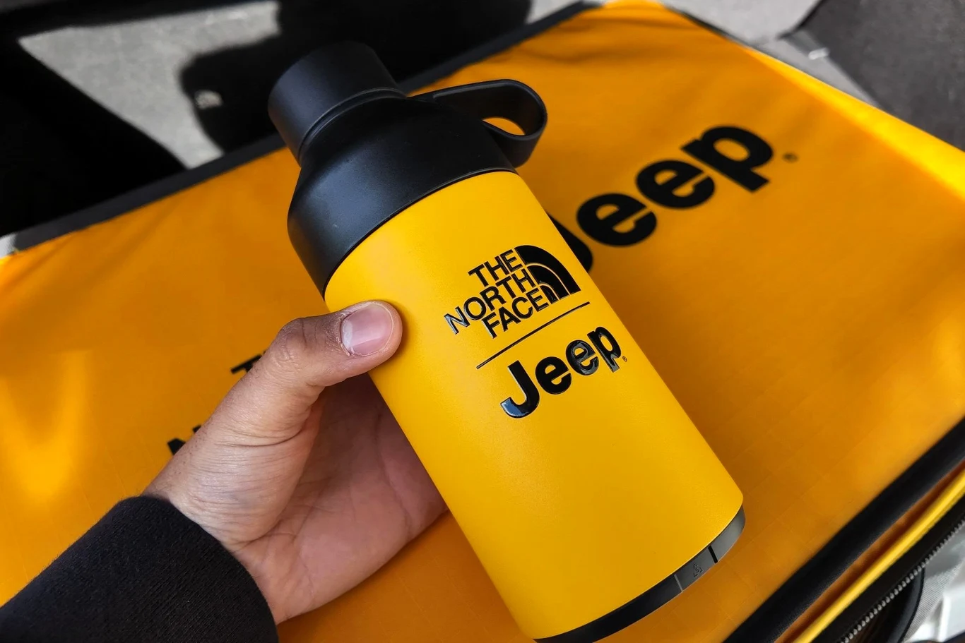 Żółta butelka termiczna z czarną zakrętką trzymana w dłoni, posiada nadrukowane logo The North Face oraz Jeep. W tle znajduje się żółty materiał również z logo Jeep.