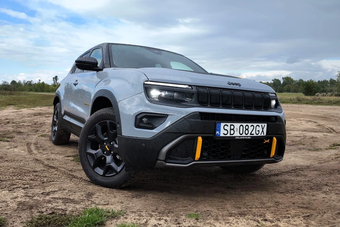 Nowoczesny SUV marki Jeep zaparkowany na piaszczystym terenie, prezentujący charakterystyczny przód samochodu z czarnym grillem, żółtymi akcentami i widoczną tablicą rejestracyjną. W tle widać pola i drzewa pod lekko zachmurzonym niebem.